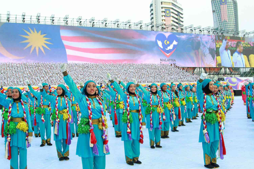A large group of Malaysian performers in matching turquoise outfits with braided pigtails and shiny tassels raise their hands in a synchronised dance against a backdrop of the Malaysian flag.