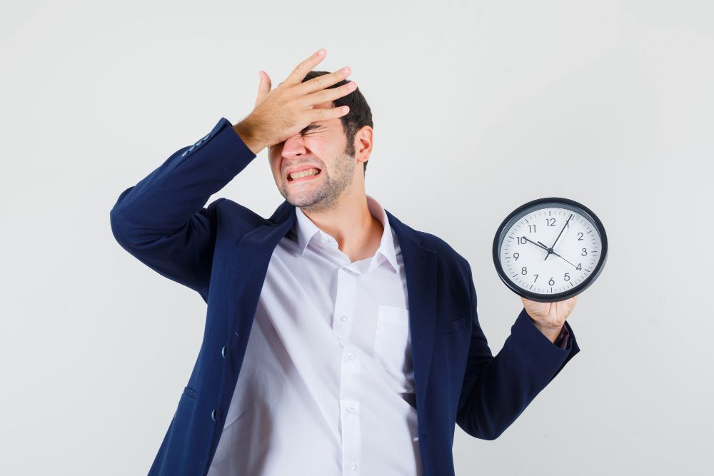 Young male holding wall clock with hand on forehead Time Delay